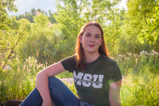 Anna, a young woman with shoulder-length red hair, sits outside with one arm resting on one knee. She wears an MSU tshirt and is surrounded by greenery and sunshine.