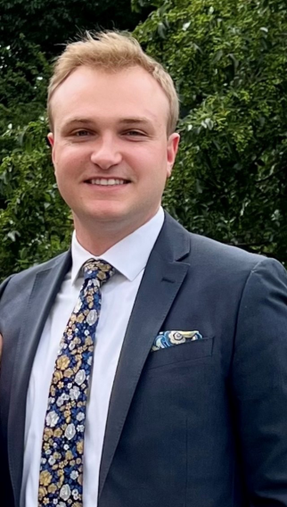 Dmytro, a young man in a suit and tie with a short haircut, smiles into the camera as he stands in front of a background of green trees.