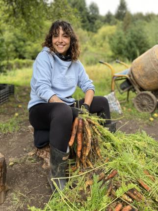 Isil, a young woman with curly, shoulder-length brown hair, sits outside on a tree sump with a bunch of freshly-picked carrots in her hands. She wears a light blue hoodie, black leggings, and black boots, and she smiles into the camera.