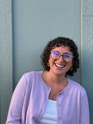 Nat, a young person with curly brown hair, is smiling widely at the camera as they stand in front of a light blue wall. They are wearing a light purple cardigan over a white tank top, round glasses, and metal hoop earrings.