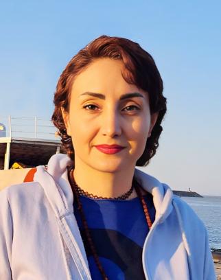 Sheida, a young woman with dark brown hair, smiles gently at the camera. She is wearing a white hoodie, blue shirt, and two necklaces, and stands in front of a body of water at sunset.