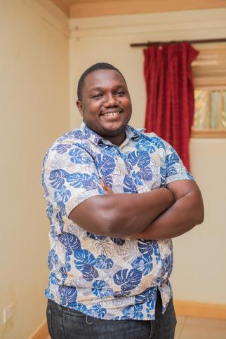 Simon, a young man wearing a blue patterned button-down t-shirt, smiles into the camera with his arms crossed over his chest. He is standing inside with a window and a red curtain in the background.