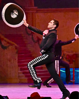 Alejandro is doing a traditional Mexican dance, wearing a traditional Mexican dancing outfit of black shirt, black pants with white detailing, and a black wide-brimmed hat with white trim. He is holding his hat in the air and his left leg is lifted off the ground as he looks upward.
