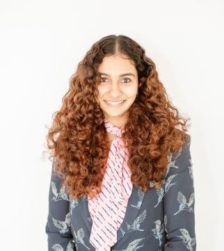 Avantika, a young woman with long, curly brown hair, smiles up at the camera as she wears a pink patterned blouse under a gray blazer embellished with birds. The background is completely white.