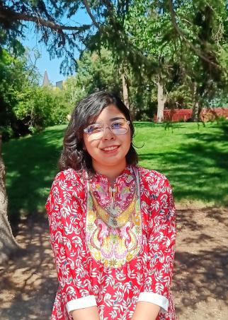 Hridi stands and smiles in partial shade on DU's campus with green grass, trees, and a blue sky behind her. Her black hair is shoulder-length, and she wears glasses and a colorful red top.