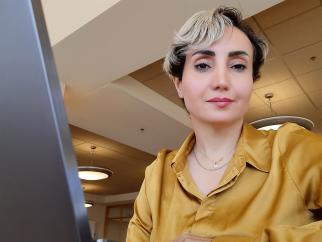 Sheida, a young woman with cropped, bleached hair, smiles gently at the camera. She wears a yellow-gold satin shirt and several dainty gold necklaces. The edge of her laptop is visible to the left of her face, and she sits in an interior space with large ceiling lights behind her.