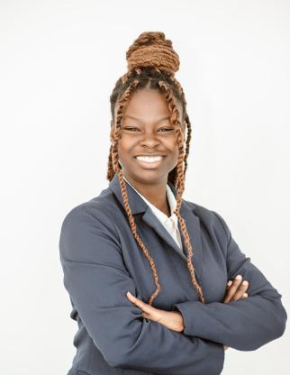 Zariah stands with her arms crossed across her chest as she smiles at the camera. She wears a black blazer and a white button-down shirt, and her hair in twisted locs sits in an updo atop her head with two face-framing twists on either side. The background is completely white.