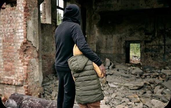 Two civilians, one of them an adult and one a child, hold each other as they look towards what appears to be a bombed building. 
