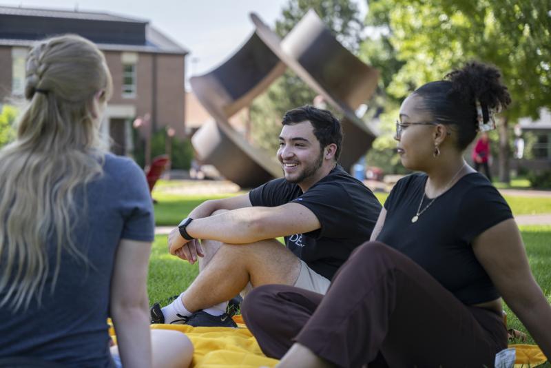 A group of three students sits and chats on the grass on campus.