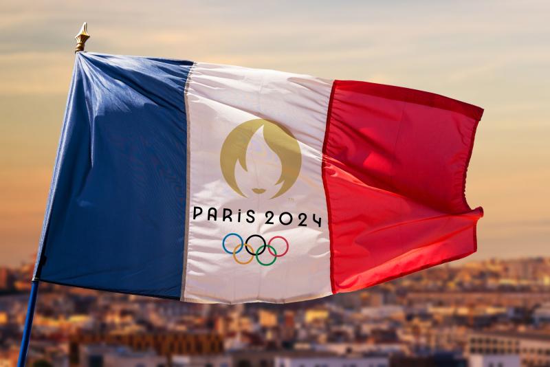 A French flag featuring the Olympic rings and the words Paris 24 flies on a flagpole over the city.