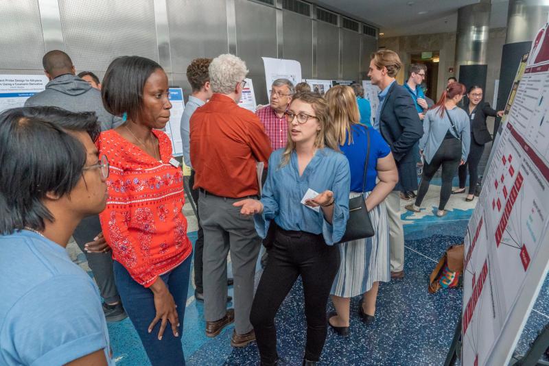 A young woman presents a trifold board research project to several onlookers.