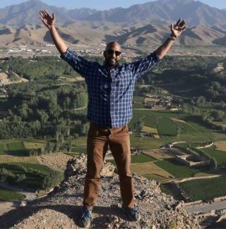 Anil Raj on a summit with arms extended in the air with mountains in the background