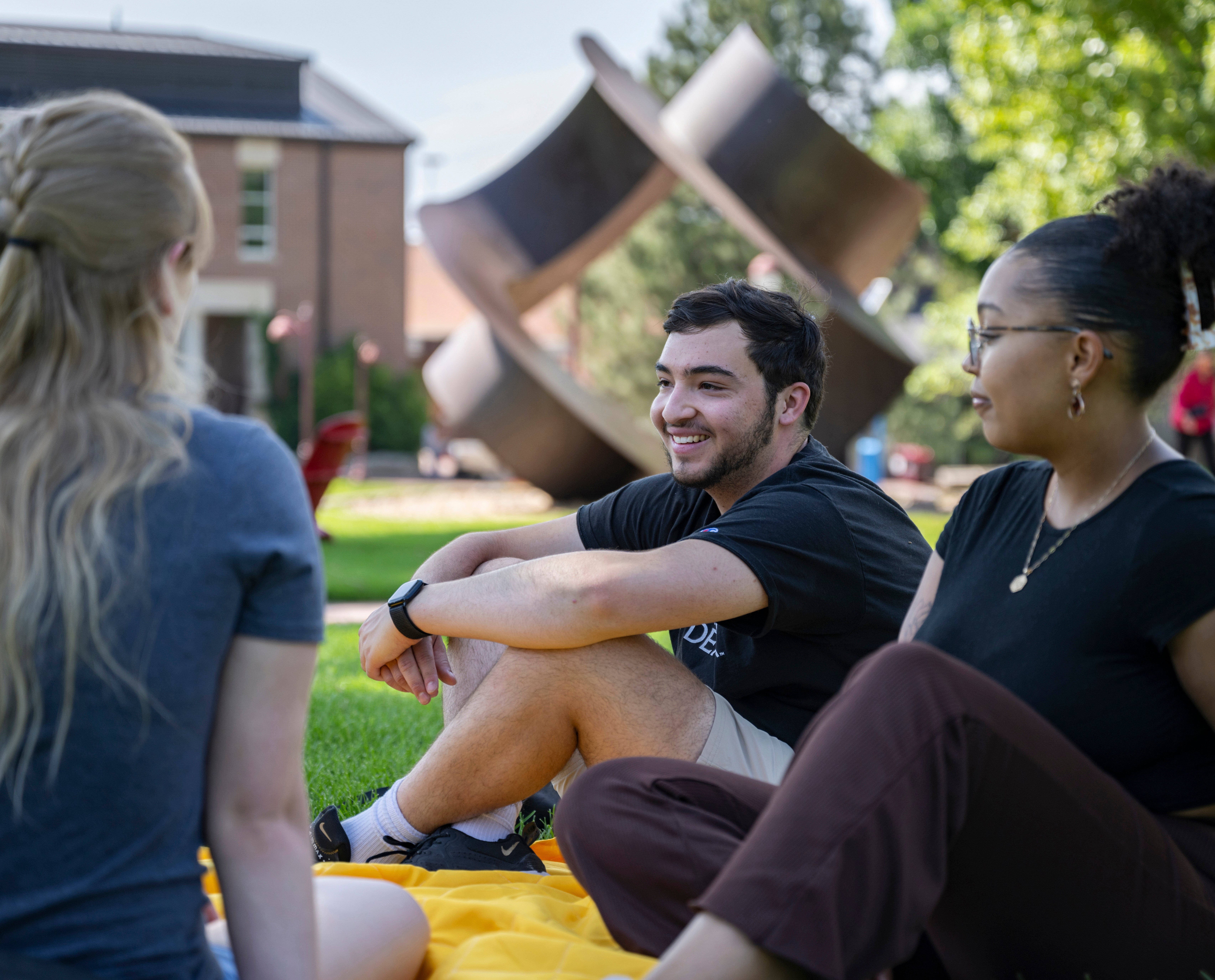 A group of three students sits and chats on the grass on campus.