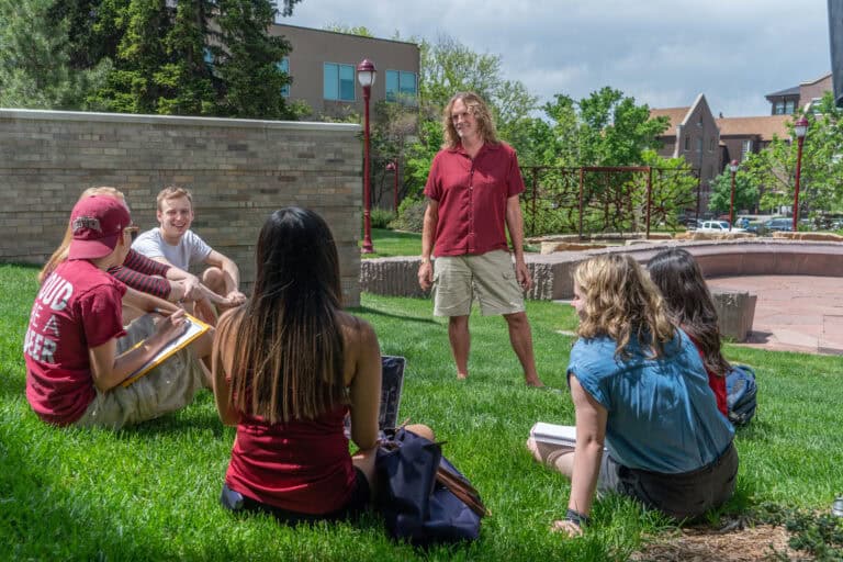 A group of students sitting on the grass outdoor being led by a standing male professor.