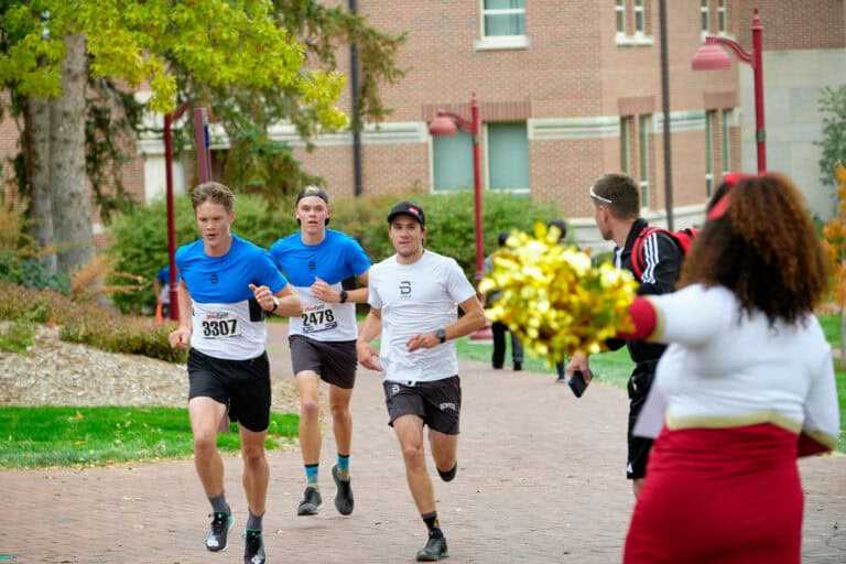 Three people participating in a 5k race with a cheerleader in the foreground.