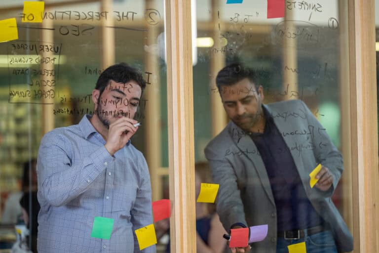 Two students writing on a glass wall filled with writing and sticky notes.