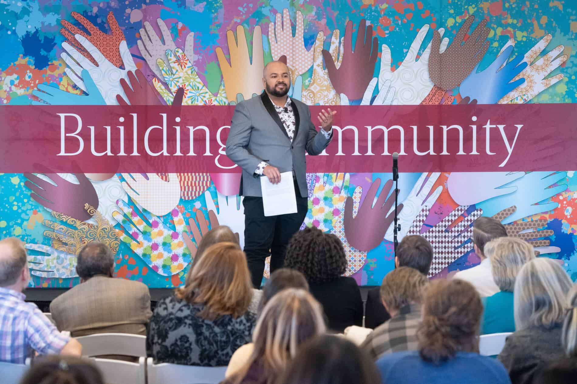 Speaker standing in front of a wall that reads "Building Community" with multi-colored hands surrounding it.